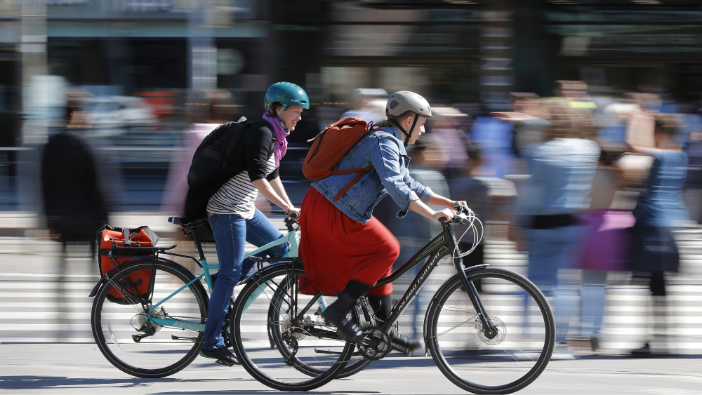 Kaksi naista pyöräilee ruuhkaisessa kaupungissa kesällä.